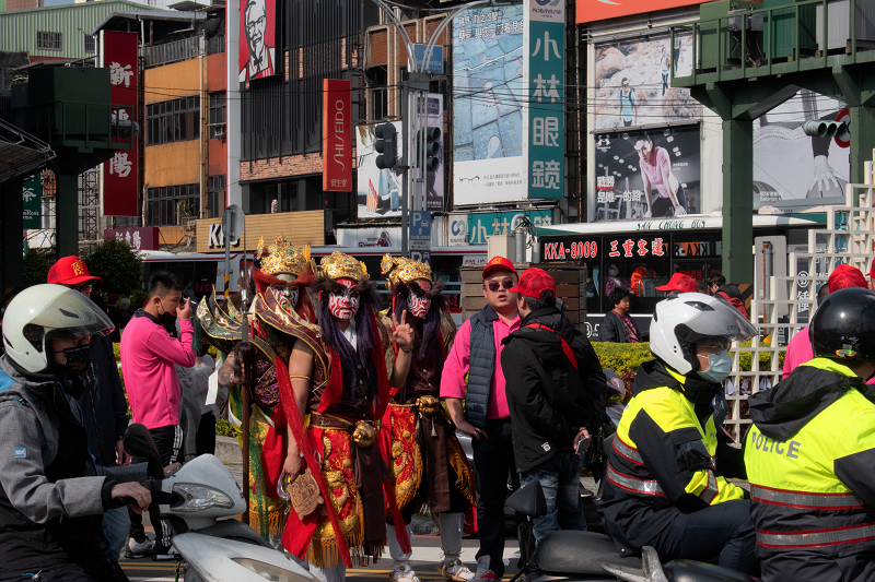 A temple procession by the Tamsui metro station. Within the procession, there are people wearing elaborate red and gold costumes while locals and police pass by on motorcycles.
