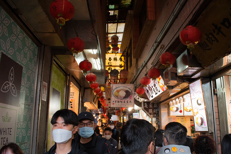 Entrance to Jiufen old street, lined with red lanterns and shops on both sides. In the middle, people are walking with masks on.