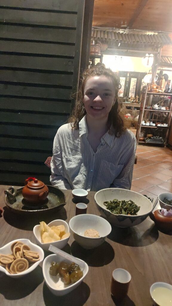 A CET Taiwan student smiling and sitting at a table in a teashop with tea snacks including dried plums, sweet potato chips, and sesame chips.