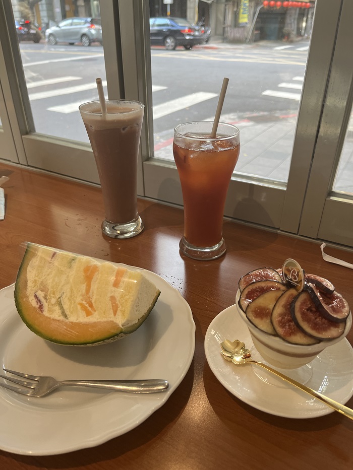 On the left plate, there is a cake inside a cantaloupe and on the right plate, there is a fig dessert. In the middle of the plates are two tall drinks.,