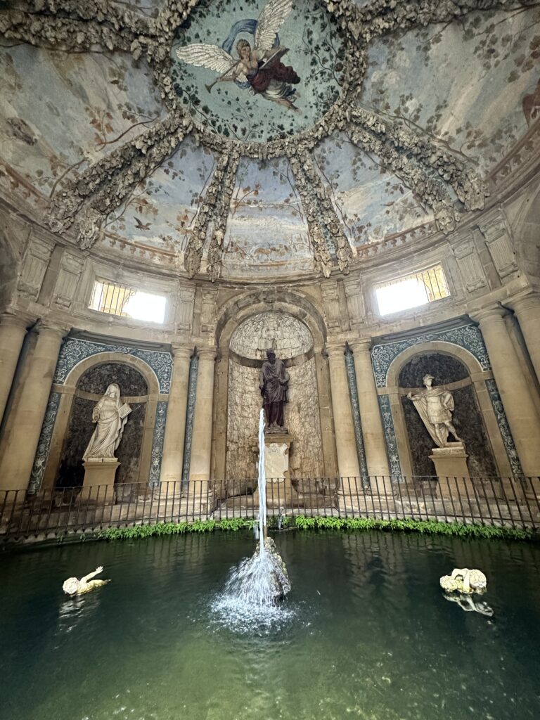 A courtyard inside Pitti Palace with marble statues, fresco paintings on the ceiling in light pastel colors, and fountain with water.
