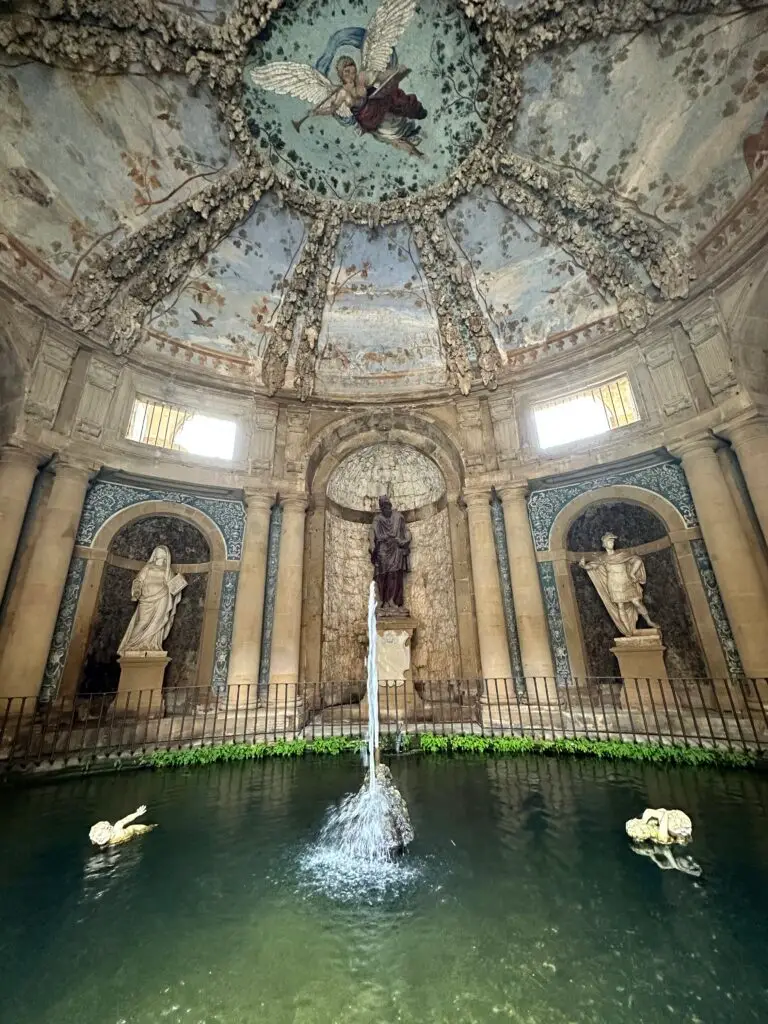 A courtyard inside Pitti Palace with marble statues, fresco paintings on the ceiling in light pastel colors, and fountain with water.