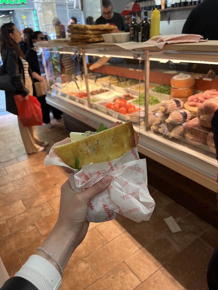 A hand holding half of a panino at the All’Antico Vinaio sandwich shop in Florence, Italy and customers standing in line to order