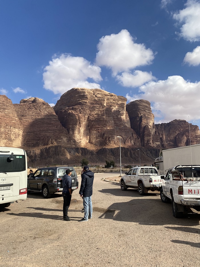 Several cars parked in a parking lot in Wadi Rum, Jordan with two people standing and looking towards the large cliffs
