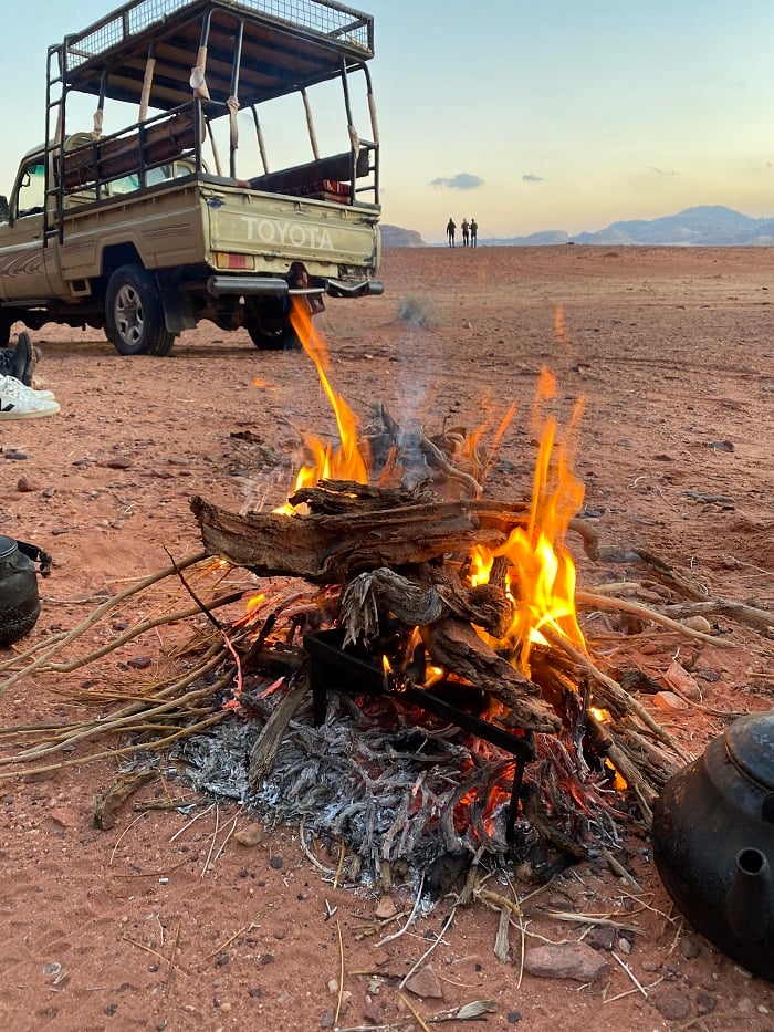 A light green Toyota truck parked to the left and a campfire on the ground in Wadi Rum, Jordan with black teapots