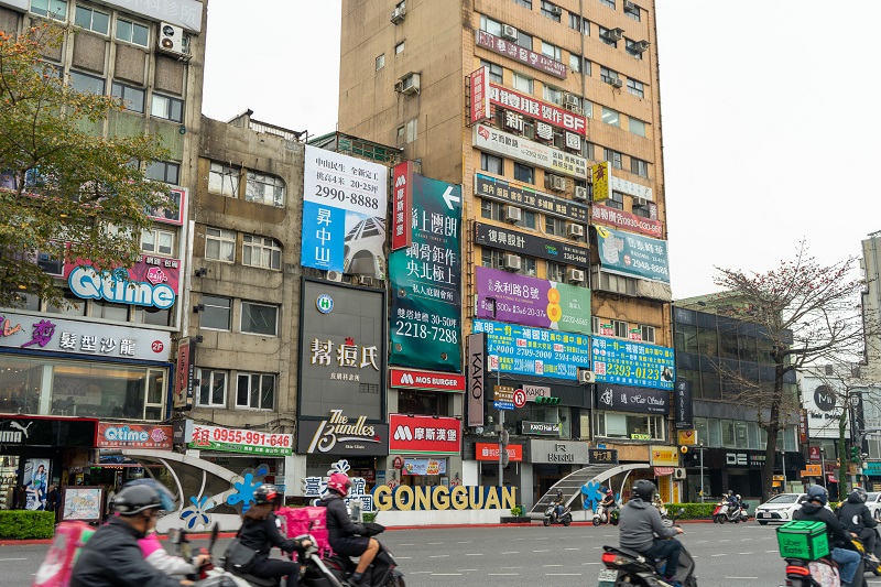 A street in Gongguan with different buildings and signs, a larger sign that reads "Gongguan" across the street and people riding motorcycles