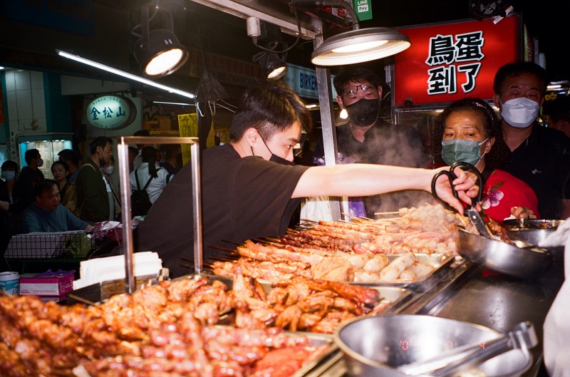 A man working behind a food vendor stand at Raohe Night Market, in front of an array of hot foods on skewers and waiting customers
