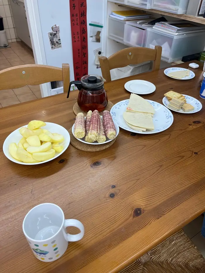 A wooden table with a pot of tea and plates with snacks like apples, corn, fruit cookies, and wafers