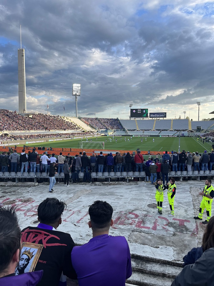 Many people in the stands and perimeters of an outdoor Fiorentina soccer game on a cloudy day