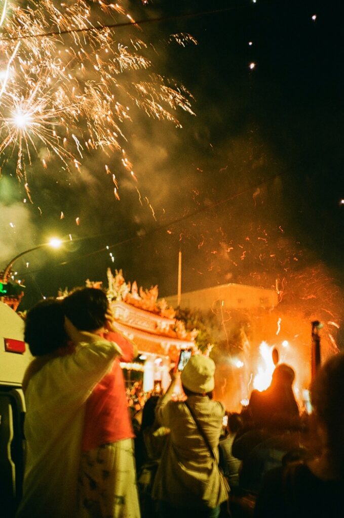 A large fire ablaze in front of Bao’an Temple, parents protecting their children’s ears, and people filming the fireworks display while the fireworks continuously going off in the background
