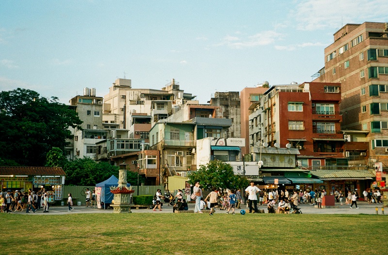 Many people including children and families strolling and playing in front of colorful buildings by the waterfront near Tamshui Old Street Plaza in Taiwan