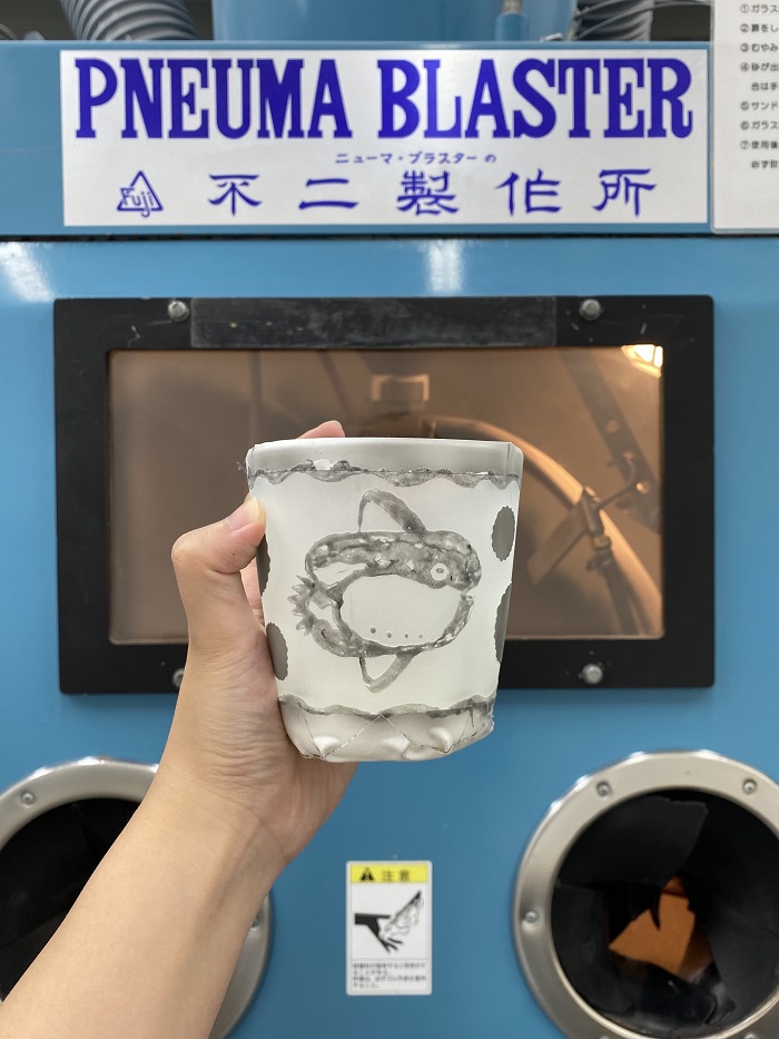 A decorated cup held in front of a machine that’s labeled “PNEUMA BLASTER” that was made in glassblowing class by an AICAD in Japan art student