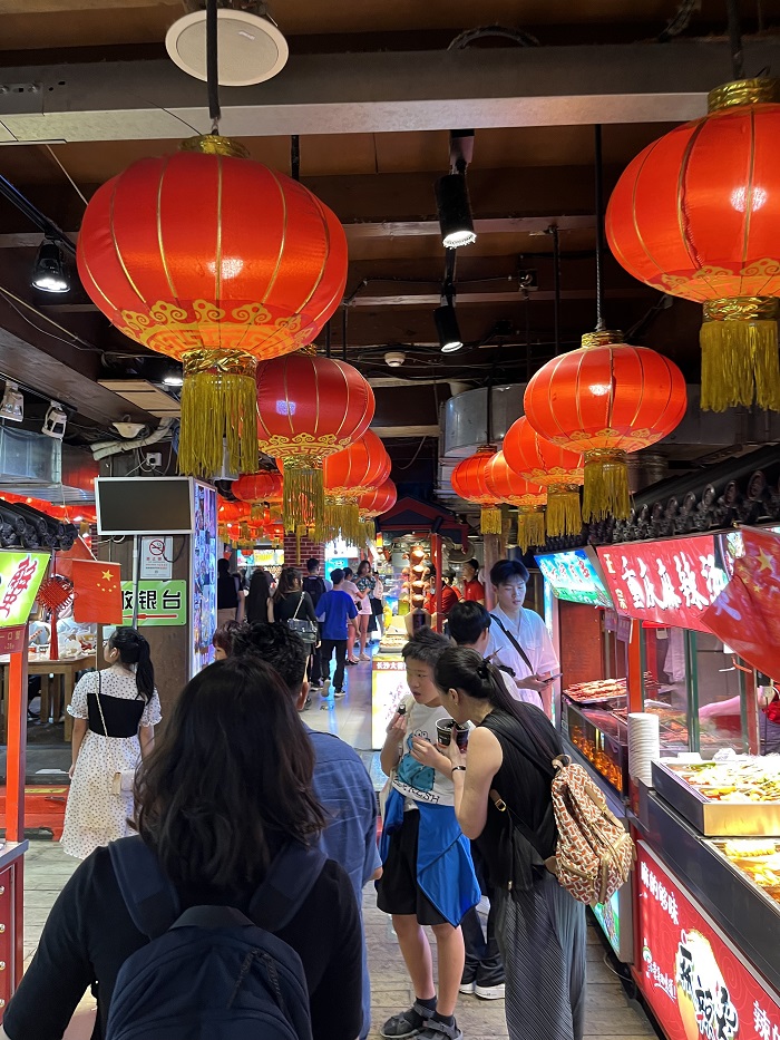 People standing in an isle with food stalls lined in Shichahai and large red lanterns hanging from the ceiling 