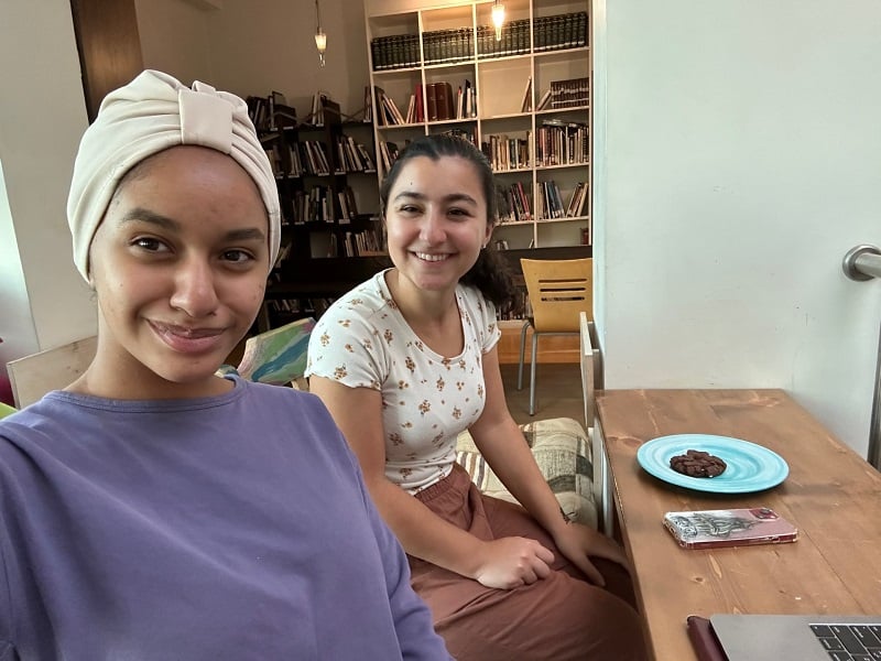Two girls smiling at the camera while sitting at a table in a café in Jordan with a bookshelf with many books in the background