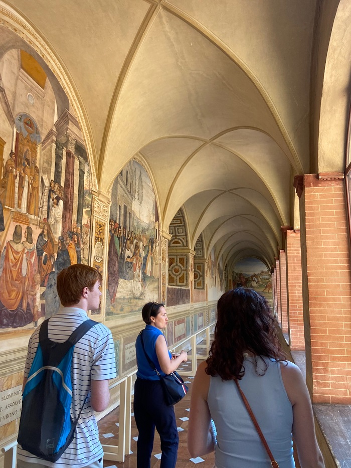 Three people walking within the Abbey of Monte Oliveto Maggiore by a wall of frescos