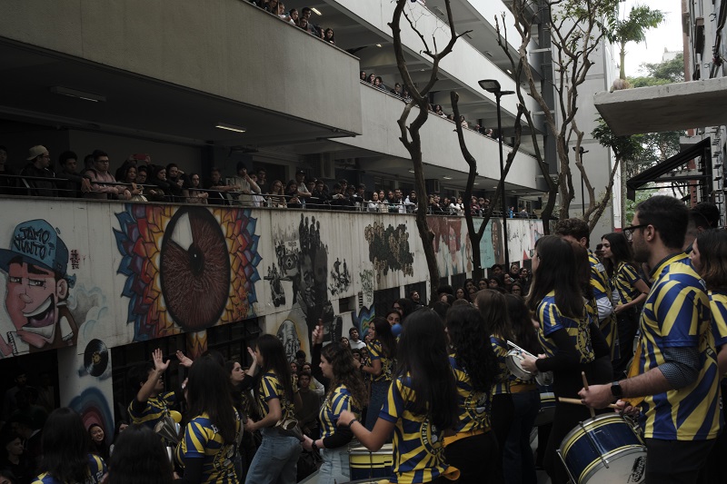 Students dressed in blue and yellow at Pontifícia Universidade Católica de São Paulo playing the drums with bystanders surrounding the performers