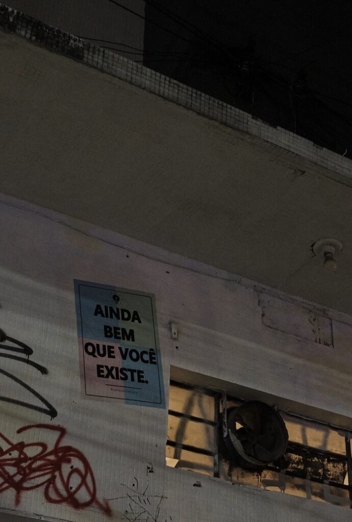 A graffiti poster glued on the side of a building in Pinheiros that reads, "Ainda bem que você existe," which translates to "I'm glad you exist."