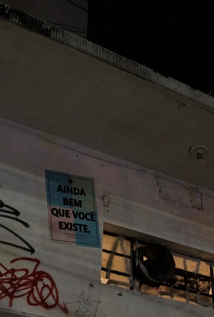 A graffiti poster glued on the side of a building in Pinheiros that reads, "Ainda bem que você existe," which translates to "I'm glad you exist."