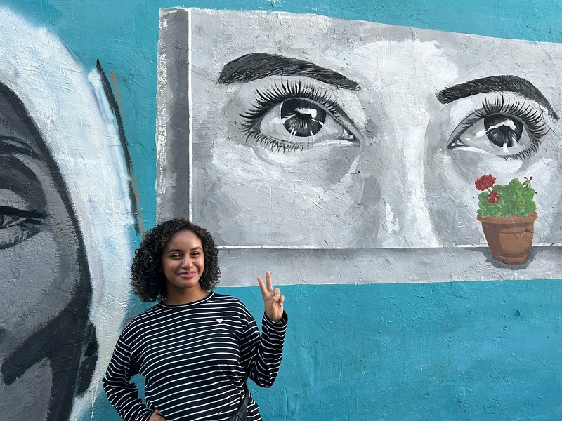 A CET Jordan student smiling and holding up a peace sign by wall graffiti on the street of a person's eyes