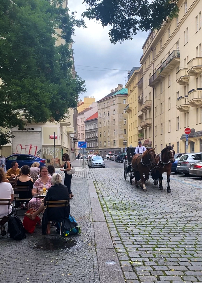 A street scene of people sitting outside, cars parked, horse and a carriage in Prague