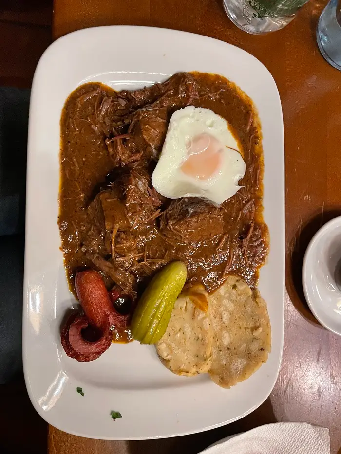 A plate of traditional Viennese Goulash including meat stew, topped off with Viennese sausage, a runny egg, and some acidic gherkins to balance it out
