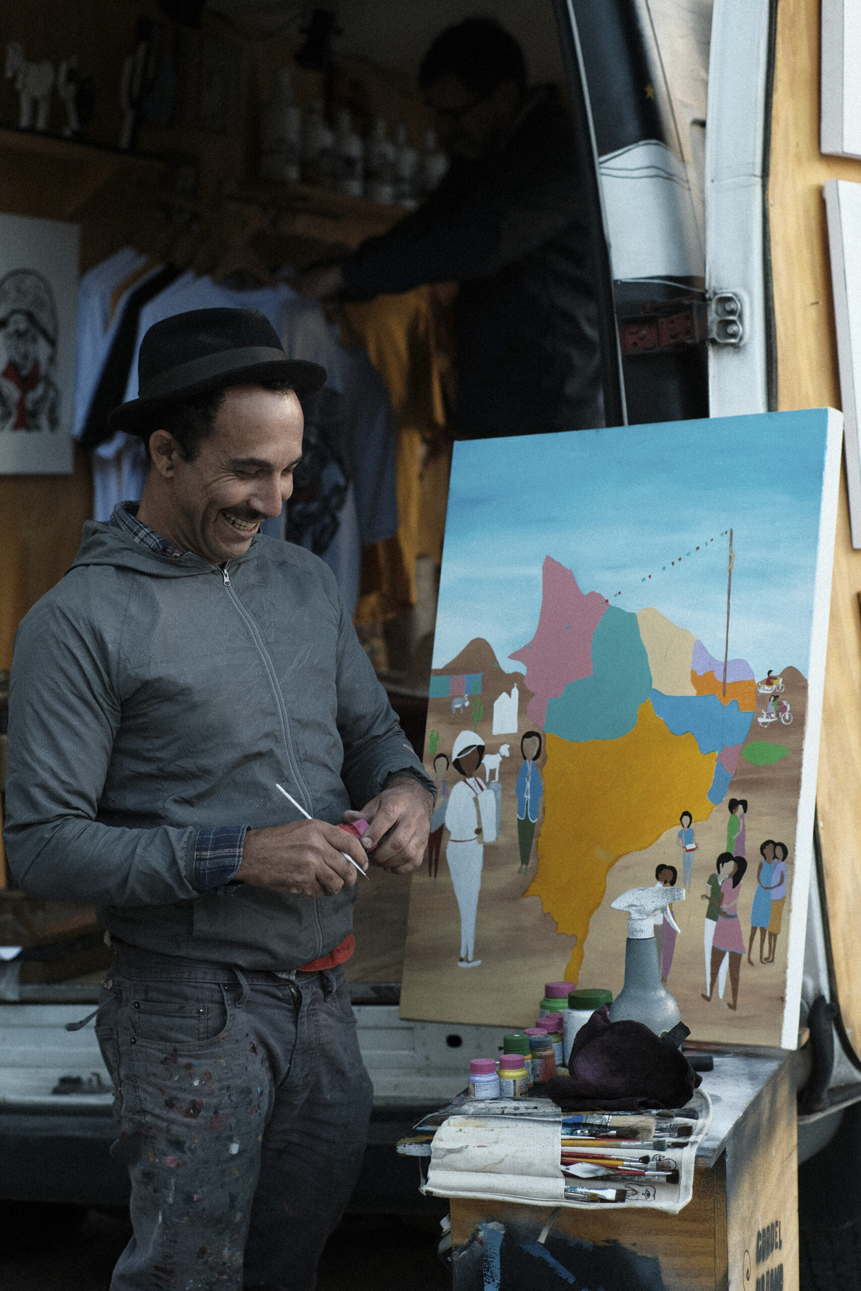 An artist in the street smiling while holding an art tool by his painting