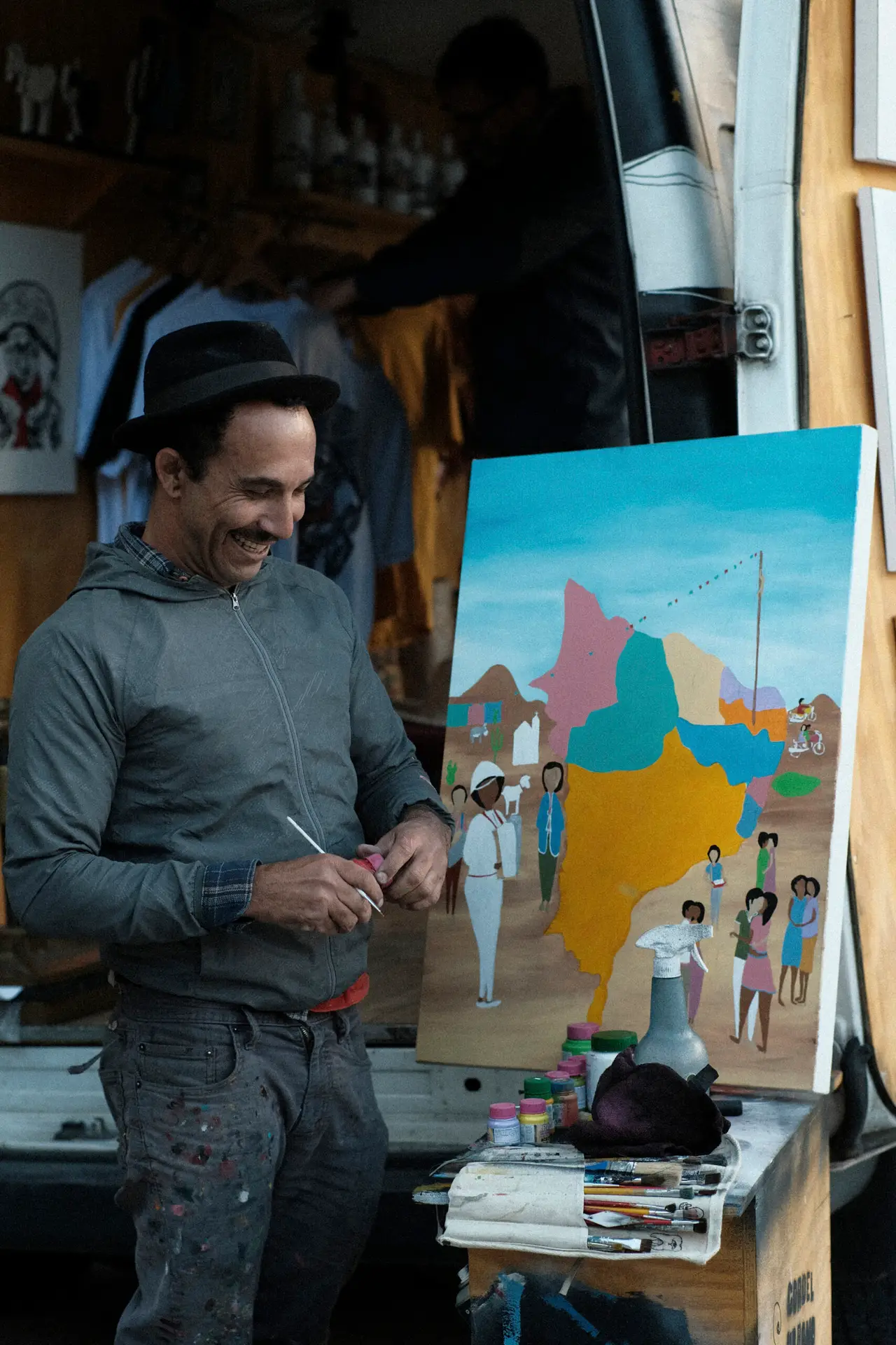An artist in the street smiling while holding an art tool by his painting