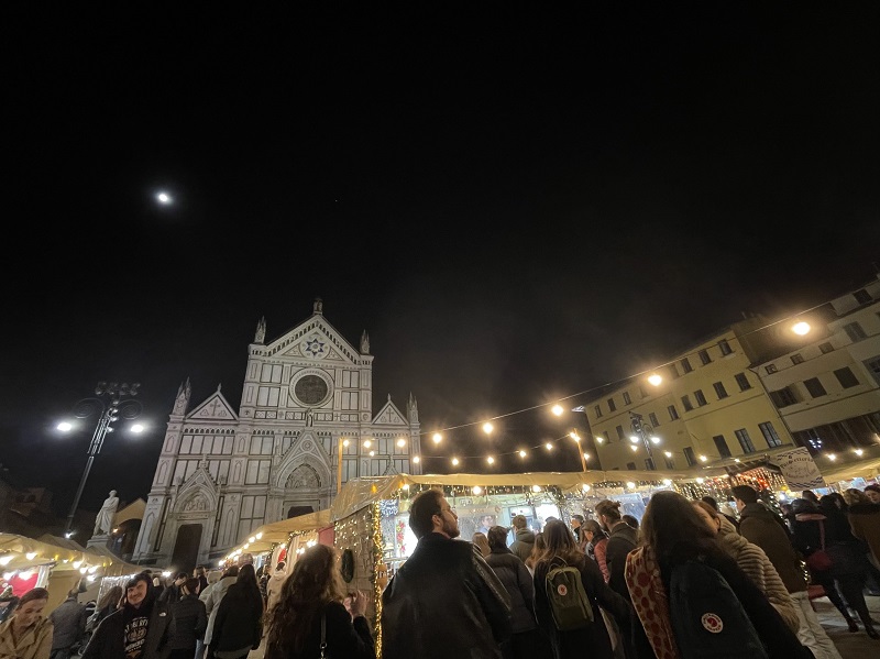 People wandering around tents under lights strung up around Santa Croce Christmas Market at night