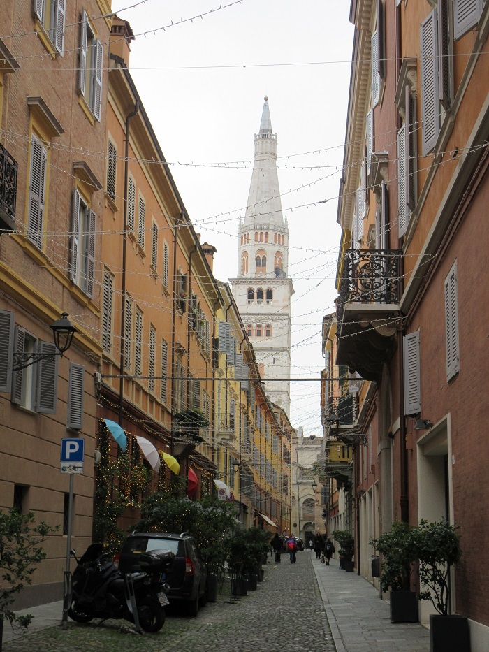 A narrow street with lights strung, people walking, and vehicles parked in Modena in Emilia-Romagna