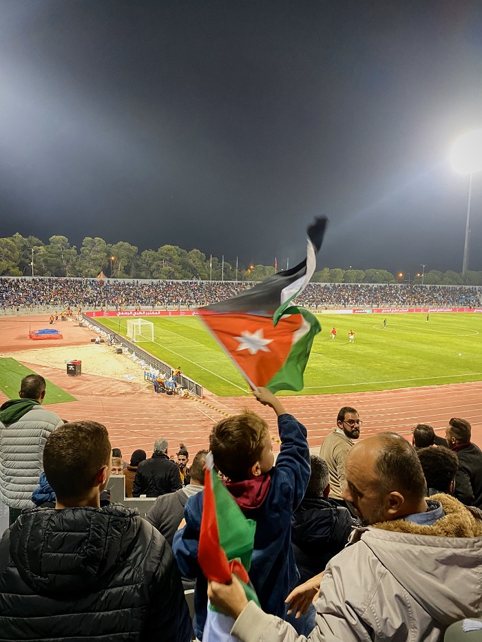People holding the Jordanian flag while at a FIFA game between Saudi Arabia and Jordan at night