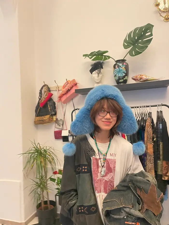 A student inside a vintage thrift store with a fuzzy hat on in front of a rack of clothes in Prague, Czech Republic