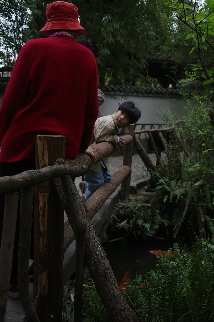 A grandmother and grandson looking at each other while leaning against a fence with koi fish swimming below by Du Fu’s Thatched Cottage
