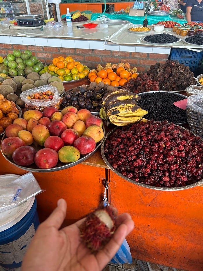 A hand holding out a lychee fruit by a large table of fresh produce in Cali, Colombia