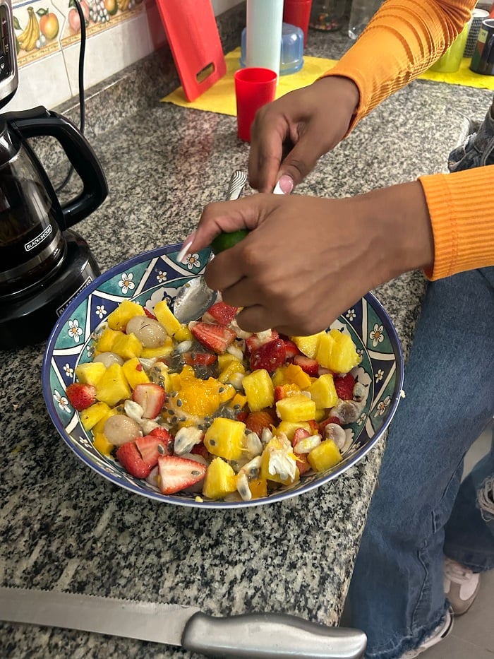 A hand squeezing out a lime on top of a bowl of fruit salad including strawberries, mangos, and more