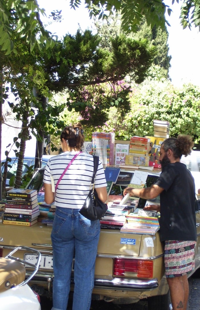 A woman looking through books with the owner on top of a car under a tree in Amman, Jordan