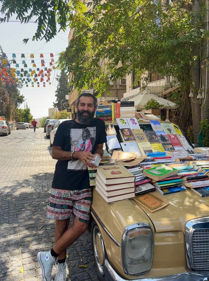 Ghaith Bahdousheh posing in front of his car covered in books while holding bookmarks in Amman, Jordan