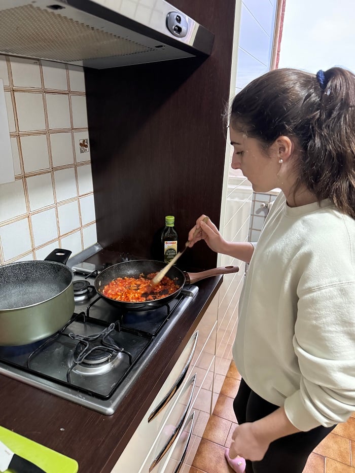 A student stirring a pan with crushed tomatoes by a pot of boiling water on the stove in a kitchen