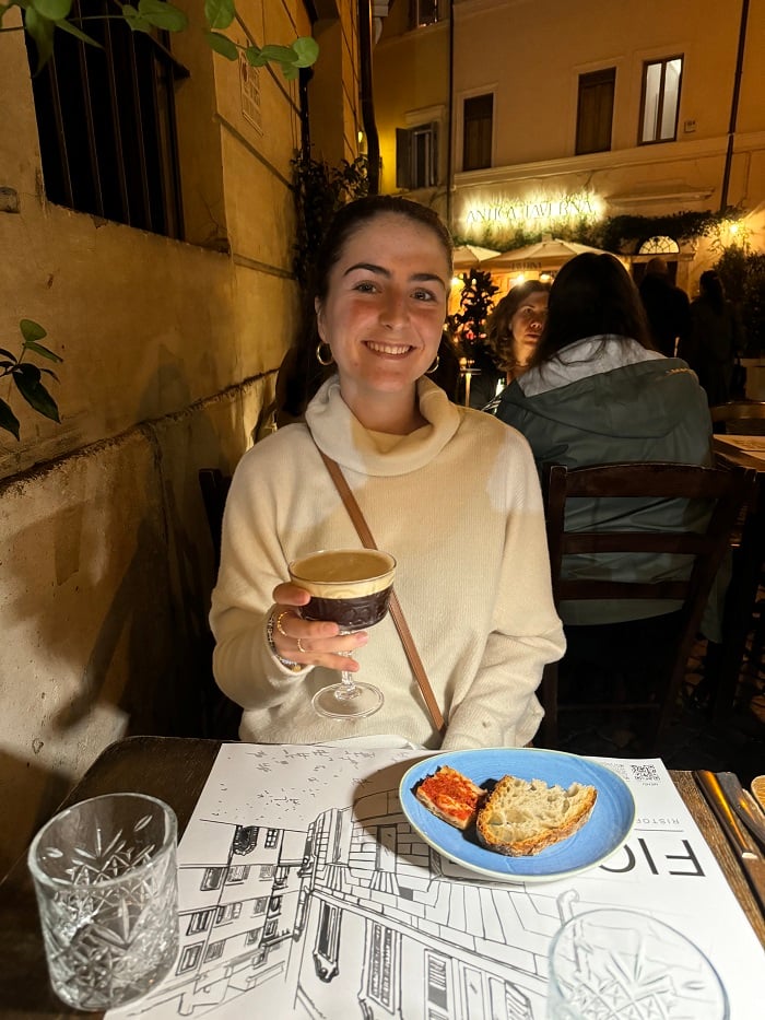 A CET Siena student holding up a drink while smiling sitting at a restaurant with a table of bread outdoors in Siena, Italy