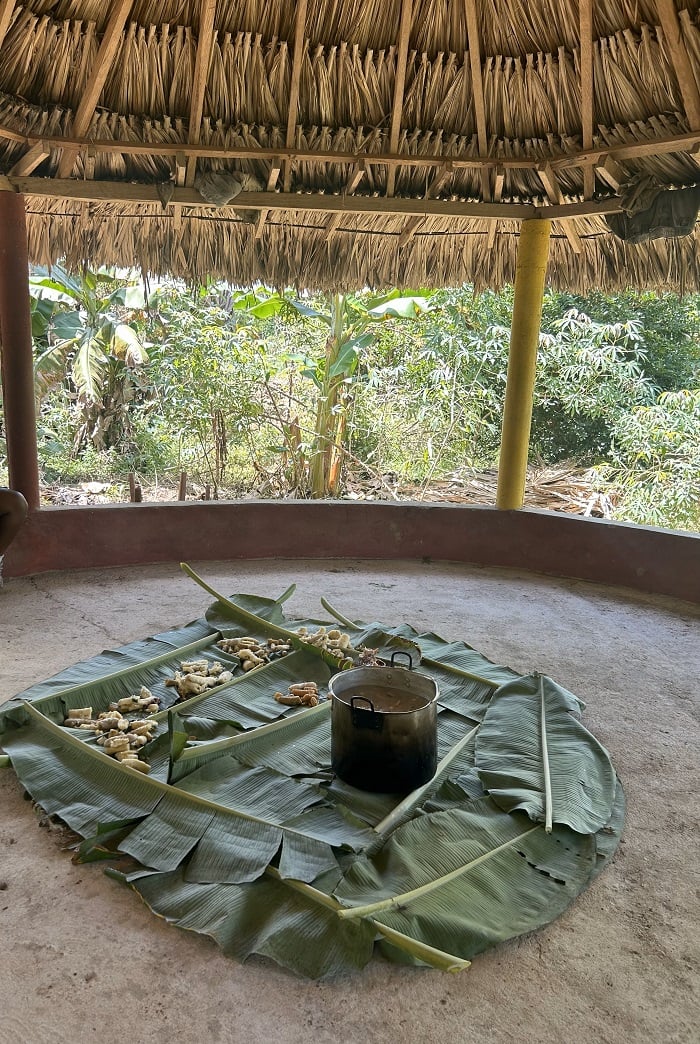 A large pot of soup placed on banana leaves on the floor under a straw roof in San Basilio de Palenque