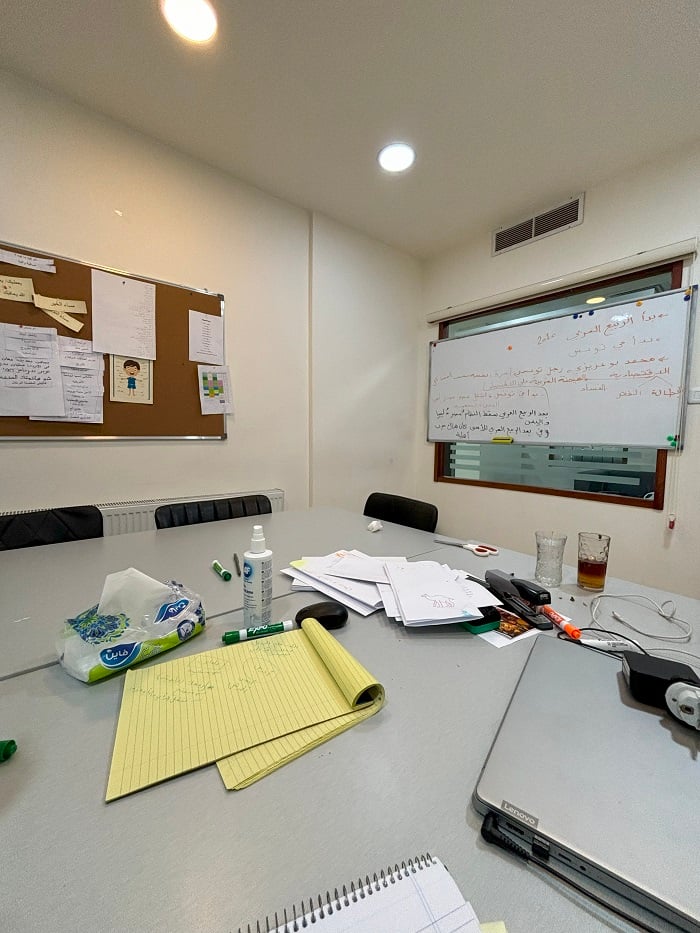 A white board with Arabic characters, bulletin board with papers, and different school supplies scattered in a classroom in Amman, Jordan