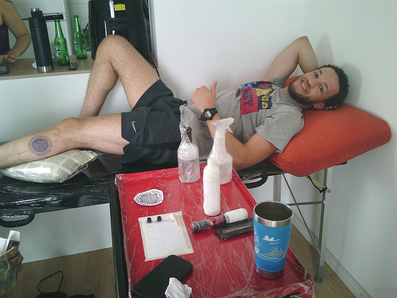 A male CET Brazil student laying down while getting a tattoo done on his lower left leg