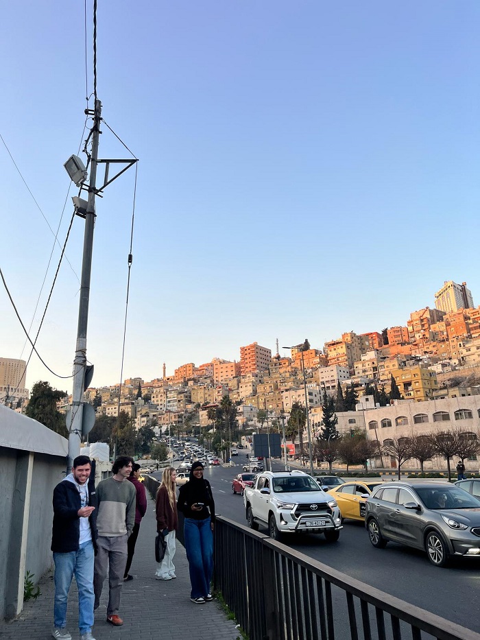 A couple of students walking along a road with cars to a local market and views of buildings in Amman behind them