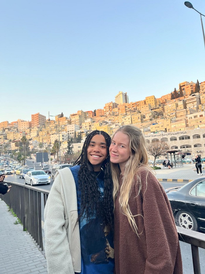 Two CET Jordan students smiling besides a road with cars to a local market and views of buildings in Amman behind them