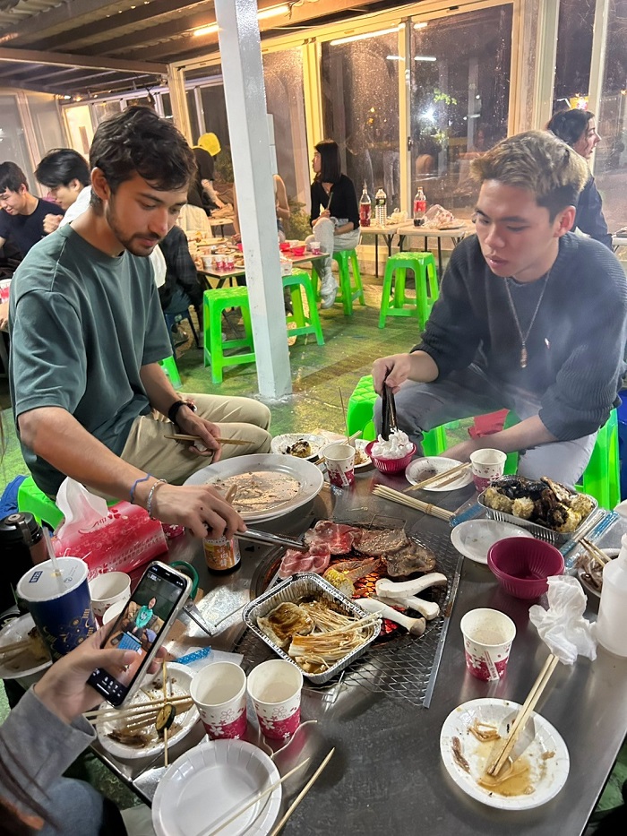 A group of people sitting around a grill eating Taiwanese-style barbeque including enoki mushrooms, tempura, lamb, shrimp
