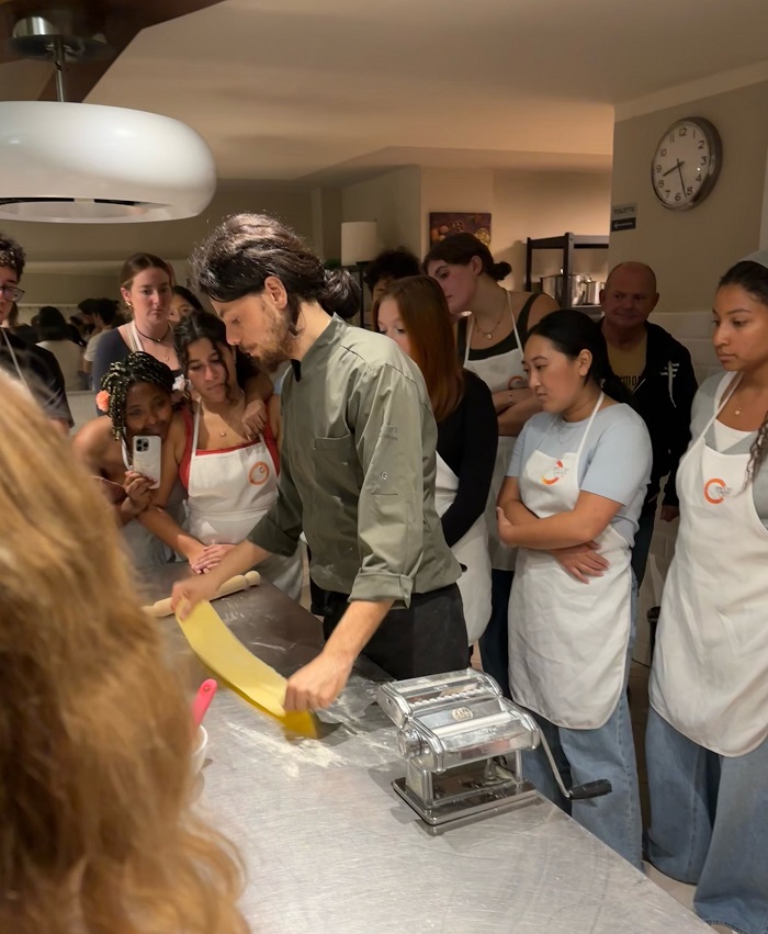 A man holding a flat piece of dough rolled out to make pasta while surrounded by many students in aprons in a kitchen