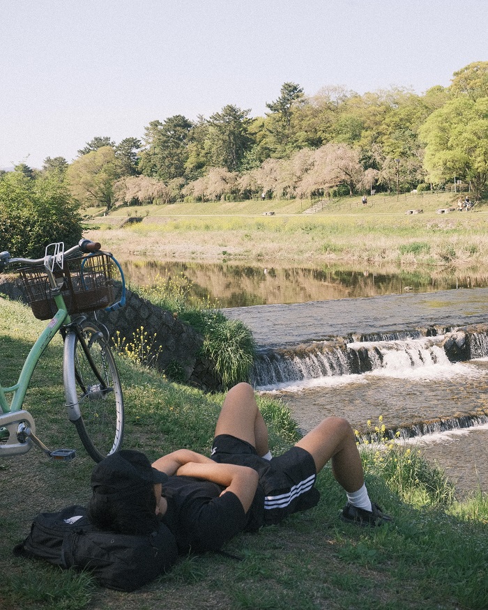 A guy laying on the grass with his bike propped up beside him by a river in Japan on a sunny day