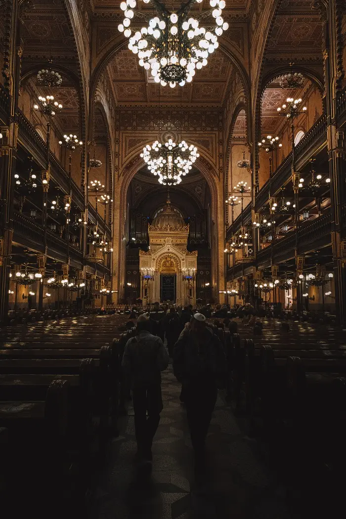 People walking down the isle with chandeliers high in the building at the Dohany Street Synagogue