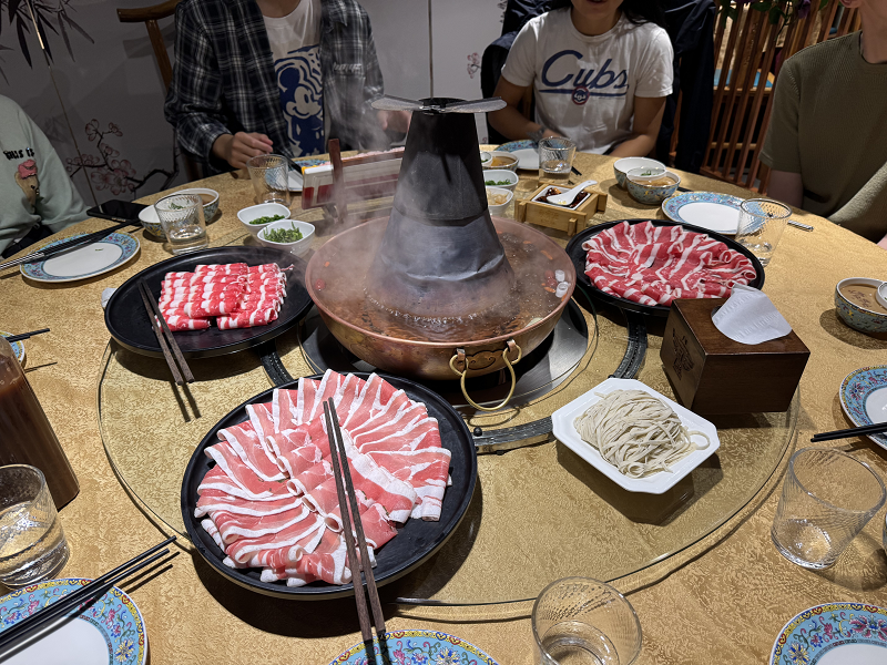 A table with different cuts of hot pot meat, a plate of noodles, and hot pot broth boiling in the middle with people sitting around the edge