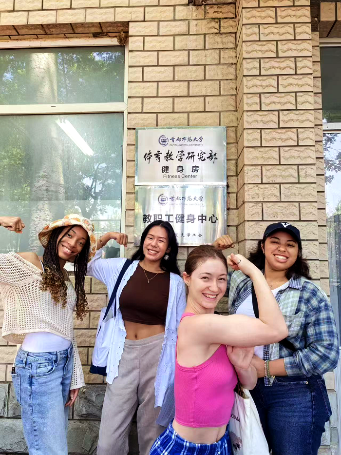 A group of four CET Beijing girls smiling and holding up their arms in front of a local university's fitness center building