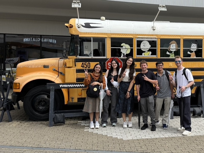A group of CET Japan students pose and flash peace signs in front of a vintage yellow school bus decorated with Peanuts characters at ExpoCity